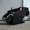 Woman relaxing in a reclined black Panasonic MAN2 5D A.I. massage chair next to a city-view window.